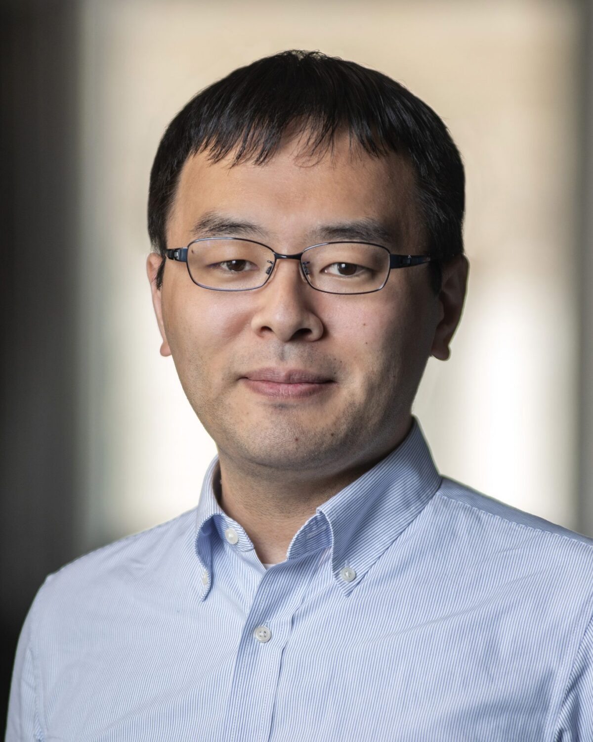 A man with short dark hair and glasses, wearing a light blue button-up shirt, stands indoors with a neutral expression. The background is softly blurred.