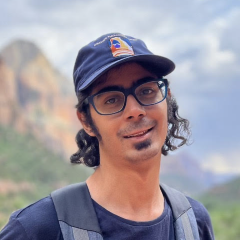 A person with glasses and a goatee, wearing a navy cap and backpack, stands outdoors with blurred mountains and sky in the background.