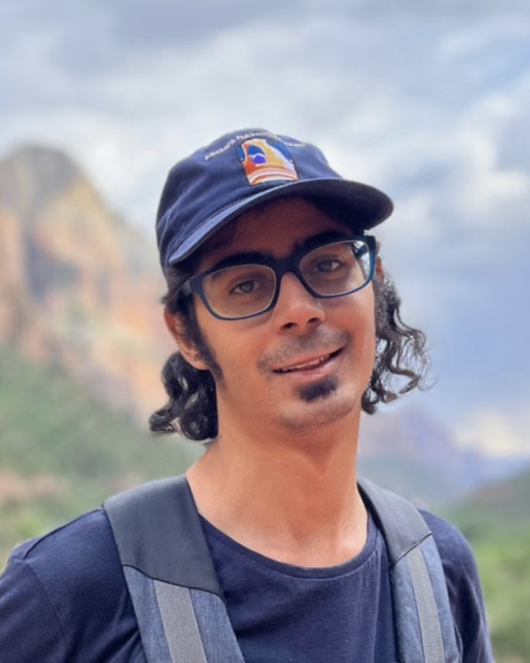 A person with glasses and a goatee, wearing a navy cap and backpack, stands outdoors with blurred mountains and sky in the background.