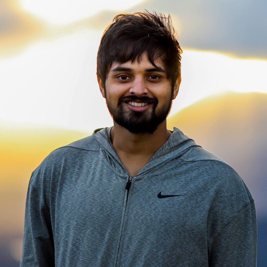A man with short dark hair and a beard, wearing a gray Nike hoodie, smiles at the camera with a bright, cloudy sky in the background.