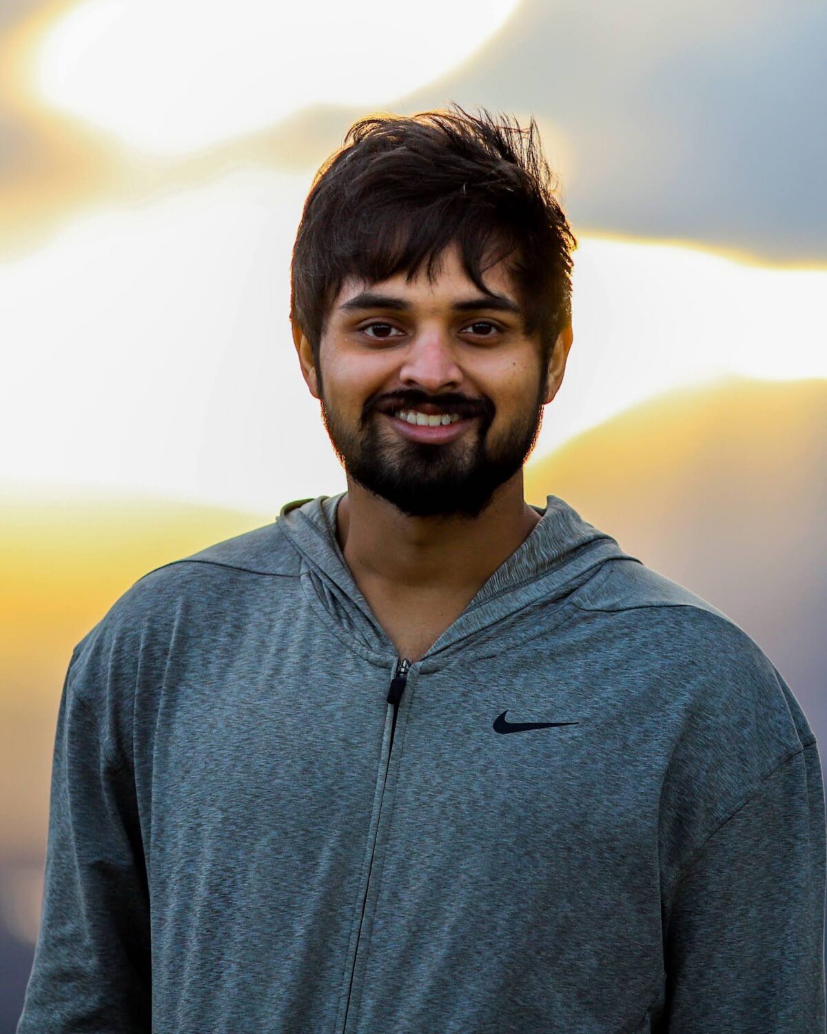 A man with short dark hair and a beard, wearing a gray Nike hoodie, smiles at the camera with a bright, cloudy sky in the background.