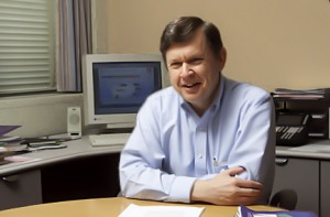 A man in a light blue shirt sits at a desk in an office, smiling. Behind him are a computer monitor, stacks of documents, and office equipment.