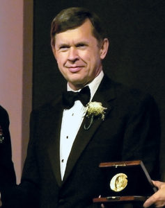 A man in a black tuxedo with a bow tie and boutonniere stands indoors, holding a plaque or box with a gold medallion.