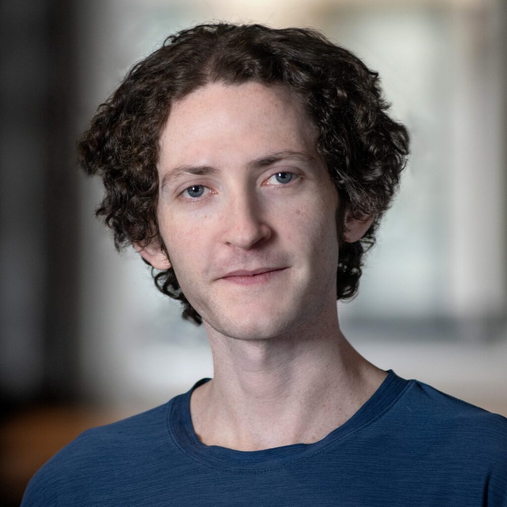A person with short, curly brown hair and light skin wears a blue shirt and stands indoors. The background is softly blurred, focusing attention on the persons face.