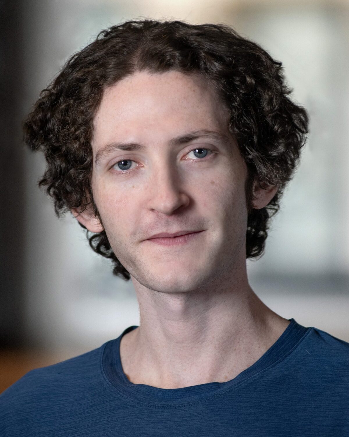 A person with short, curly brown hair and light skin wears a blue shirt and stands indoors. The background is softly blurred, focusing attention on the persons face.