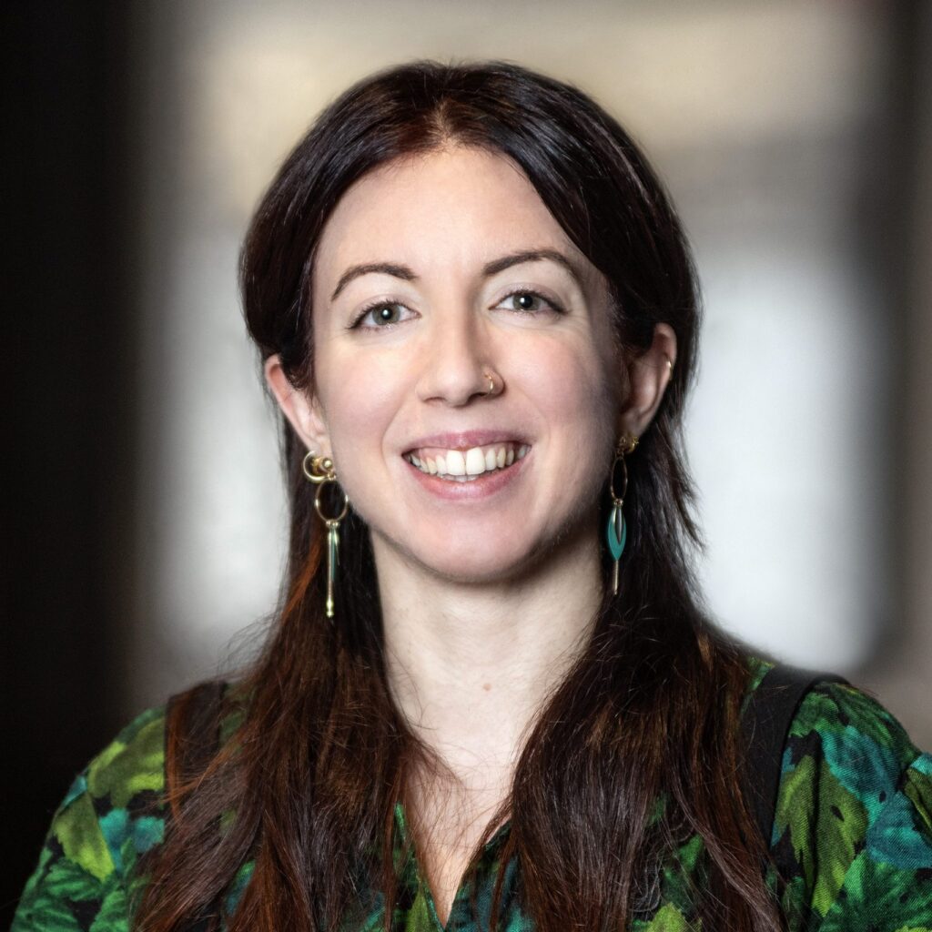 A woman with long brown hair, wearing green earrings and a green patterned top, smiles at the camera against a softly blurred background.