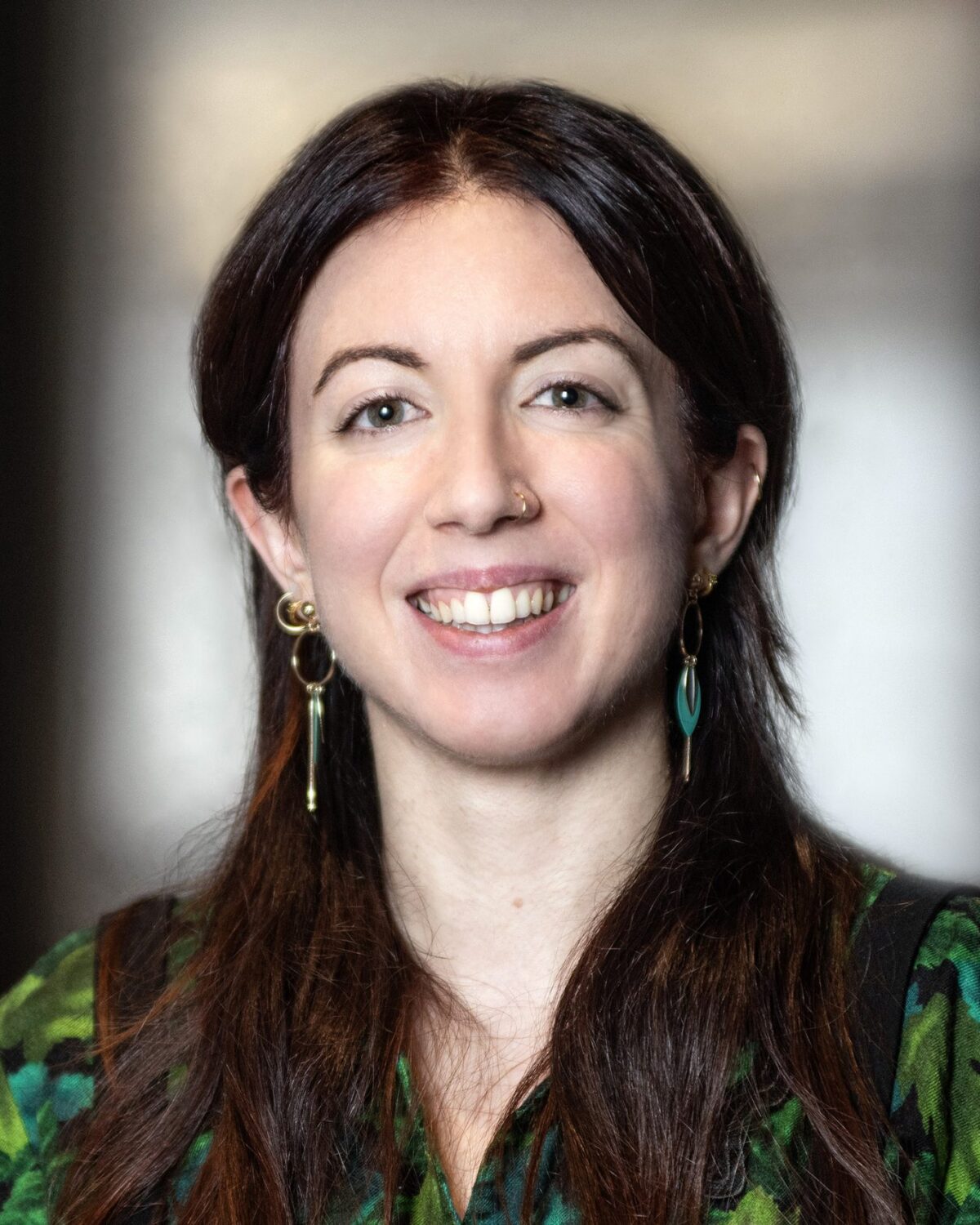 A woman with long brown hair, wearing green earrings and a green patterned top, smiles at the camera against a softly blurred background.