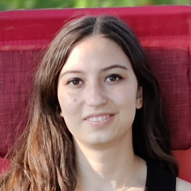 A young woman with long brown hair smiles softly, sitting in front of a red textured background outdoors.