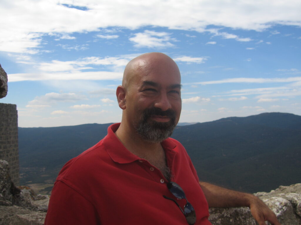 A man with a bald head and beard, wearing a red polo shirt and sunglasses, stands outdoors with hills and a cloudy sky in the background.