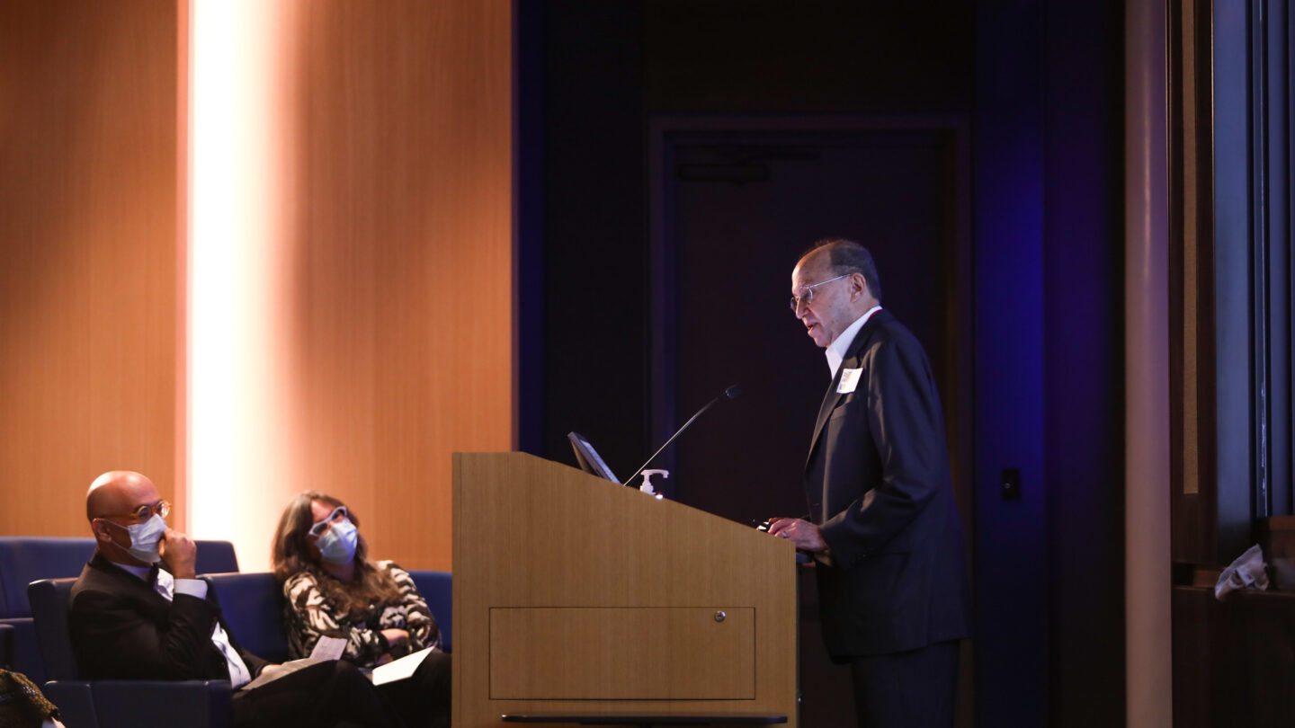 A photograph of a man speaking at a wooden podium as two people wearing masks sit in auditorium seats.