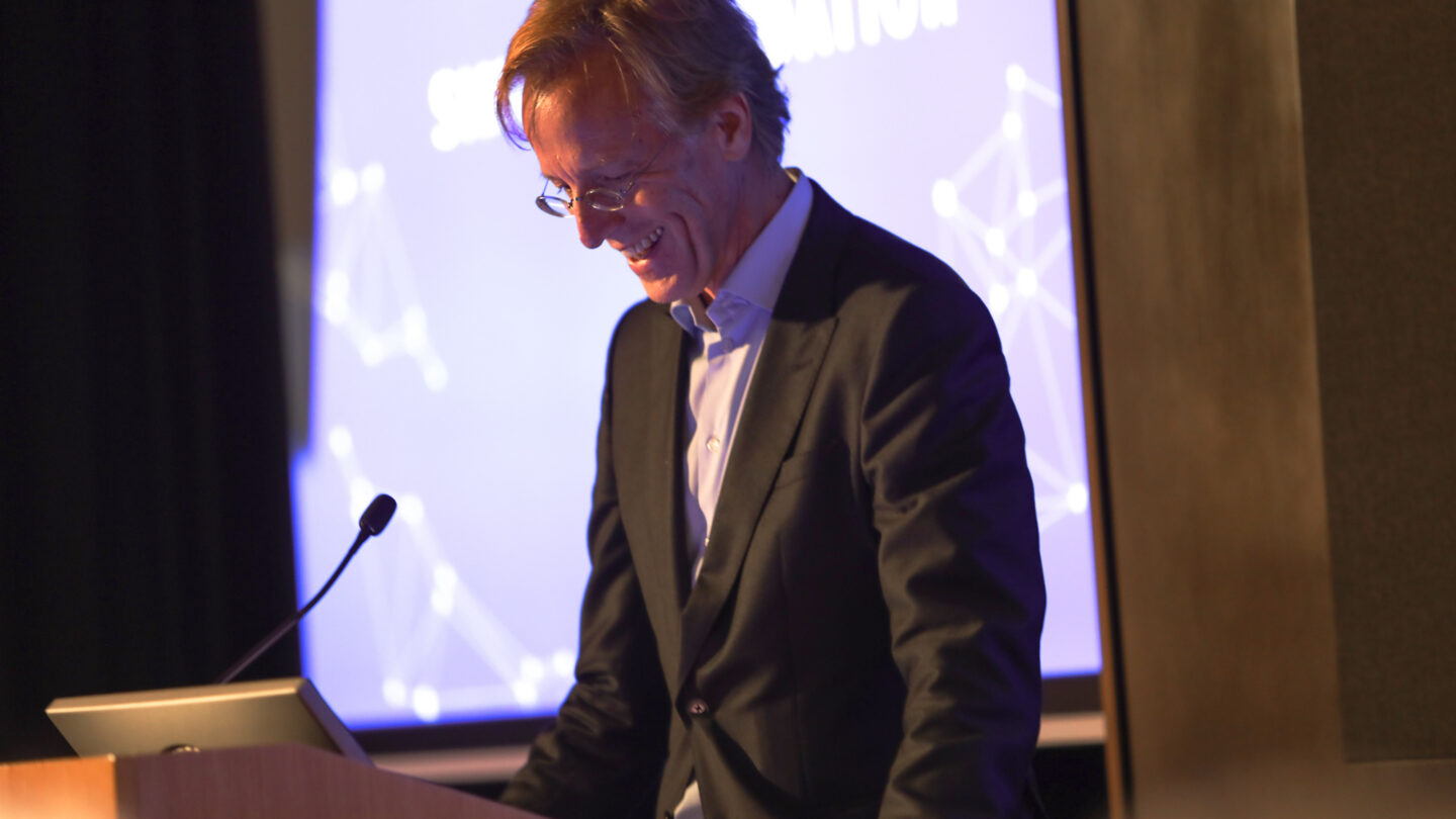 A photograph of a person speaking at a wooden podium and standing beside a screen with a slide projected onto it.
