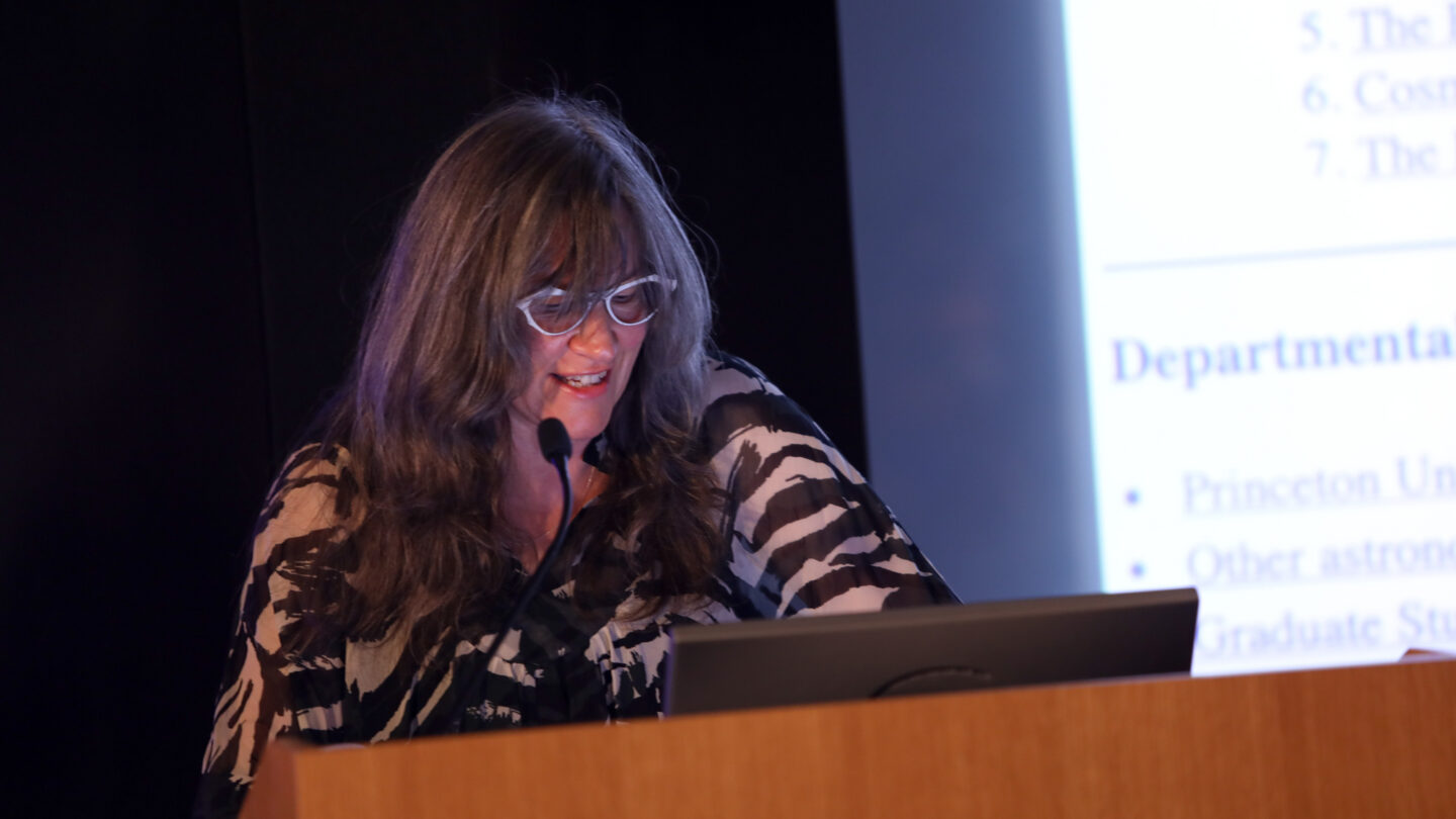 A photograph of a person speaking at a wooden podium and standing beside a screen with a slide projected onto it.