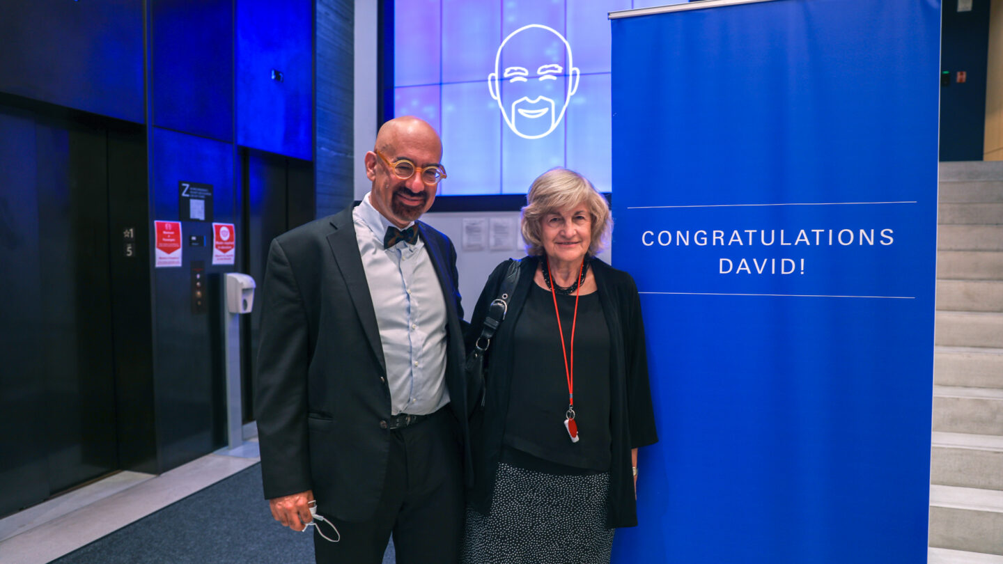 A photograph of two people smiling and facing towards the camera as they stand beside a banner that reads 