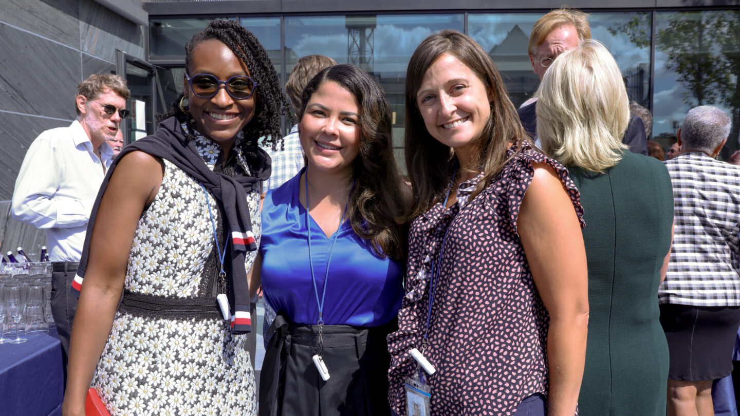 A photograph of three women smiling towards a camera as they stand in an outdoor space.