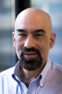 A middle-aged person with a bald head, dark eyebrows, and a trimmed dark beard and mustache, wearing a light blue collared shirt, looking directly at the camera with a neutral expression.