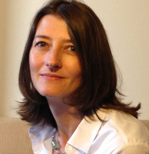 A woman with shoulder-length brown hair wearing a white collared shirt and a necklace sits on a light-colored couch, smiling softly at the camera.