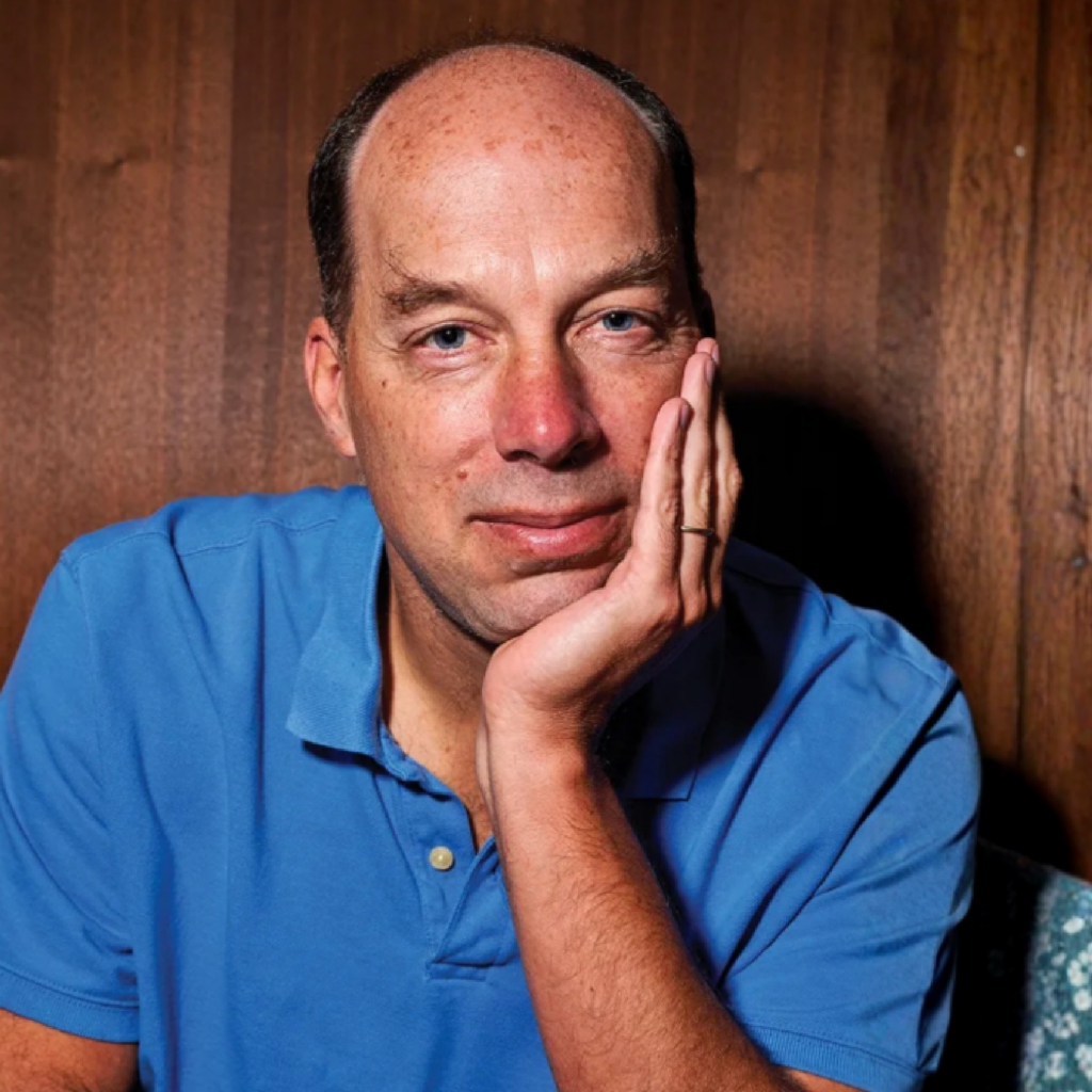A man in a blue polo shirt sits with his hand resting on his cheek, looking at the camera. He is in front of a wooden wall and appears thoughtful.