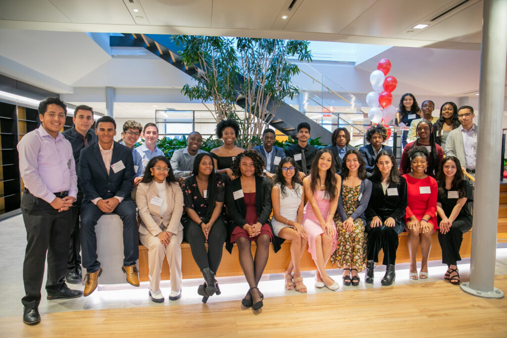 A group of young adults dressed in business or formal attire pose together indoors, smiling at the camera. Red and white balloons are visible in the background.
