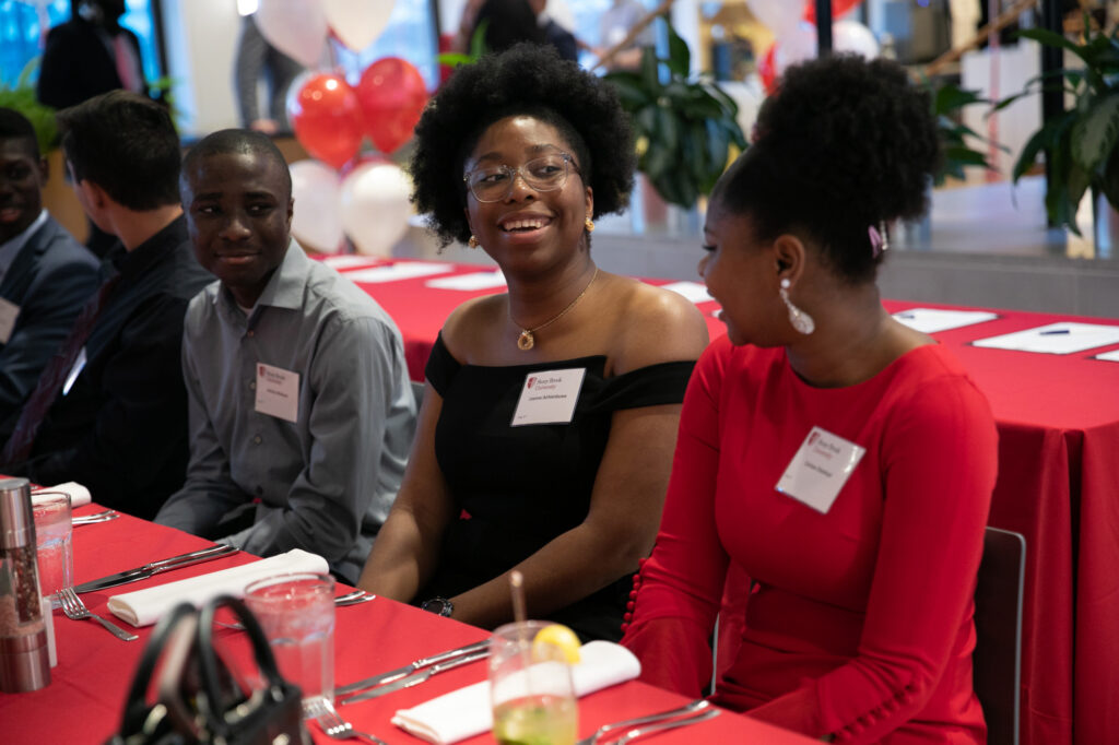 Four people sit at a red-draped table set for a formal event, smiling and talking. Red and white balloons and plants decorate the background, and everyone wears nametags.