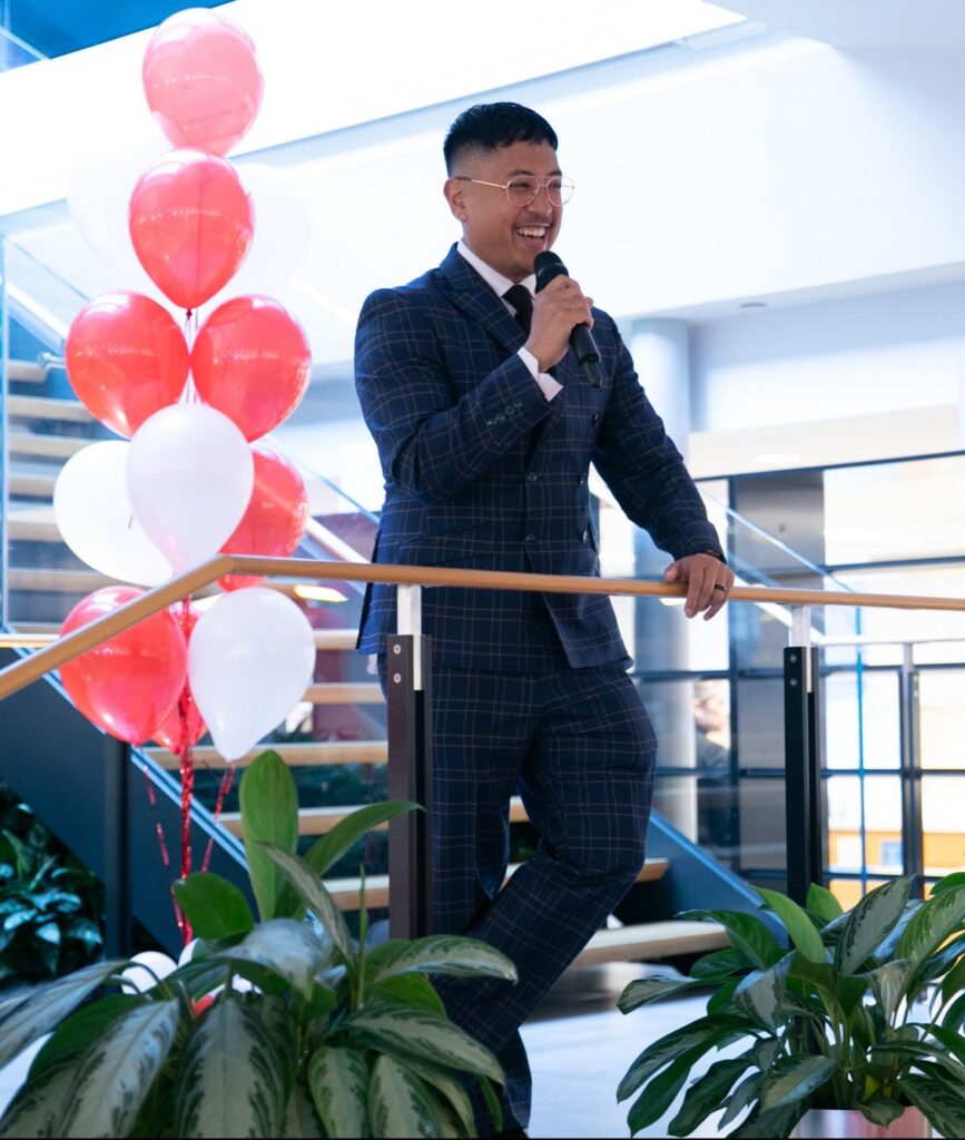 A person in a plaid suit holds a microphone and smiles while speaking on a staircase next to red and white balloons, with green plants in the foreground.
