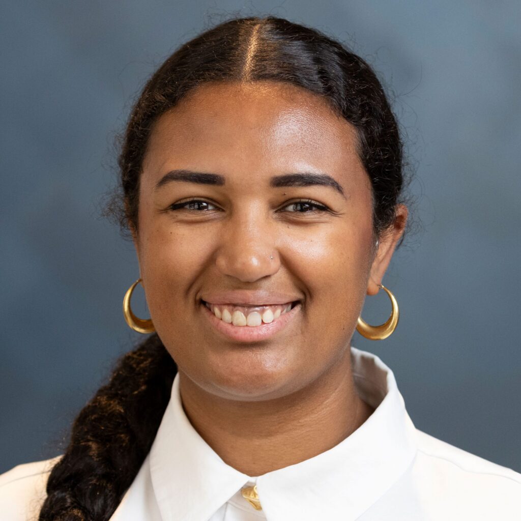 A woman with dark curly hair in a low ponytail, wearing gold hoop earrings and a white collared shirt, smiles at the camera against a blue background.