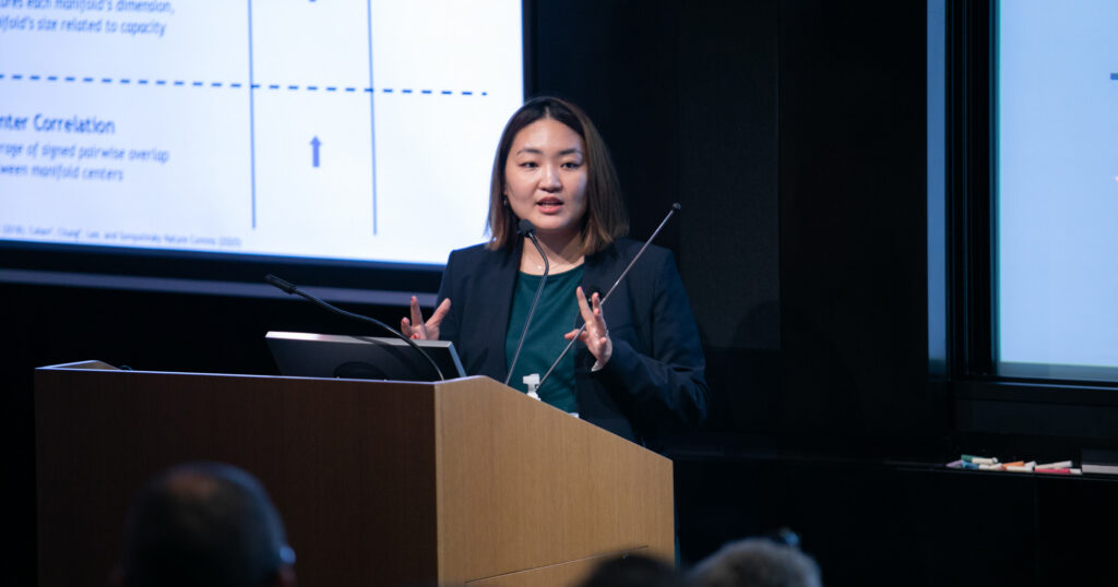 A woman stands at a podium, speaking and gesturing with her hands during a presentation. A projection screen with graphs and text is visible beside her.