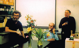 Three men are in an office, two sit at a table with flowers, while one stands holding a drink. There are shelves with folders, a whiteboard, and a bottle of champagne on the table.