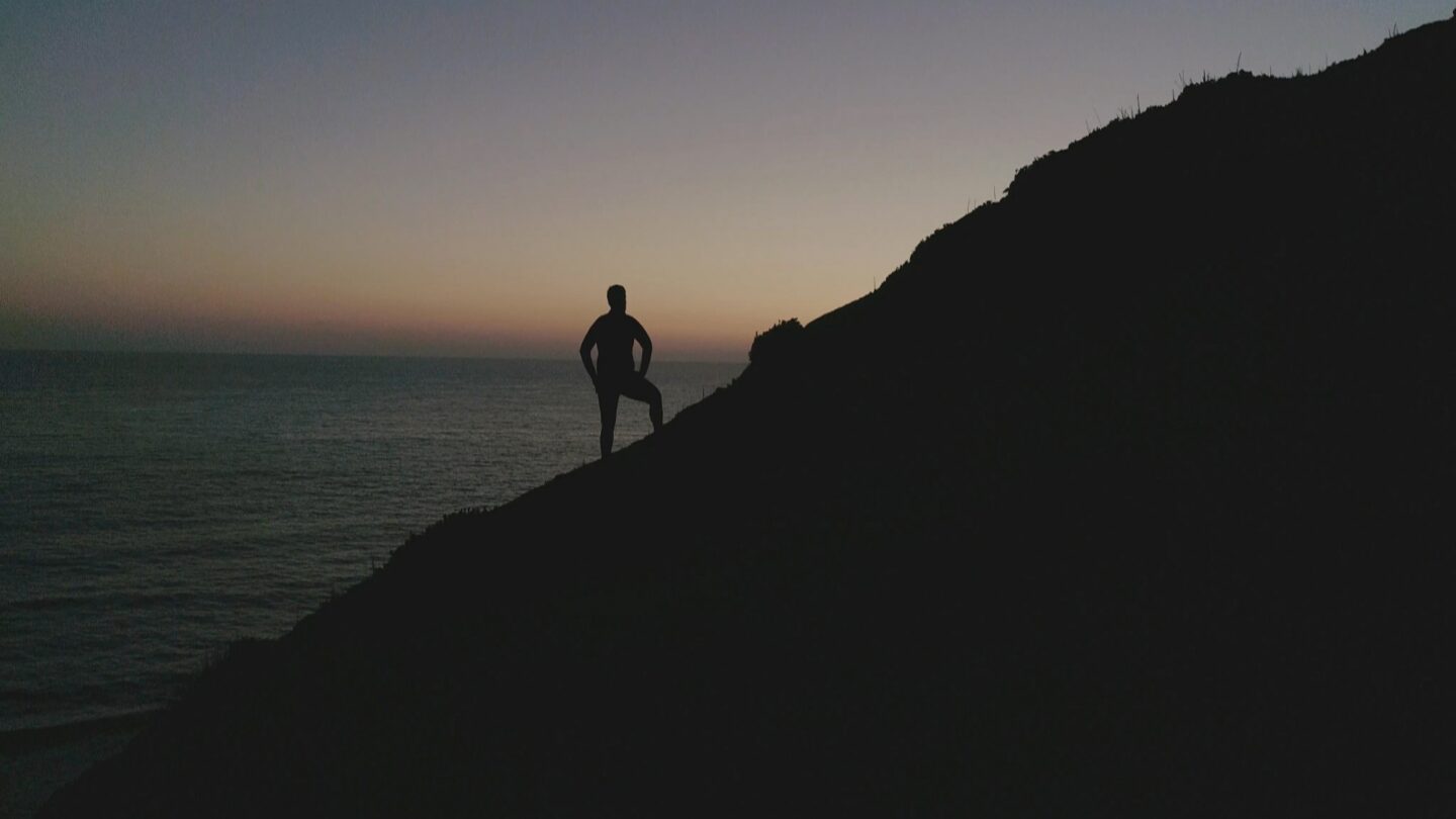 Man on a hill, backlit by a sunset on an ocean