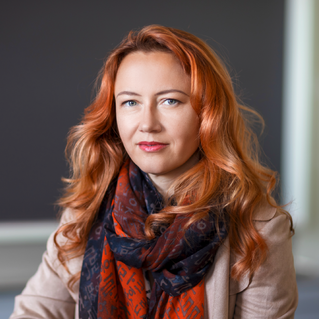 A woman with long red hair wearing a patterned scarf and beige jacket sits in front of a blurred dark background, looking calmly at the camera.