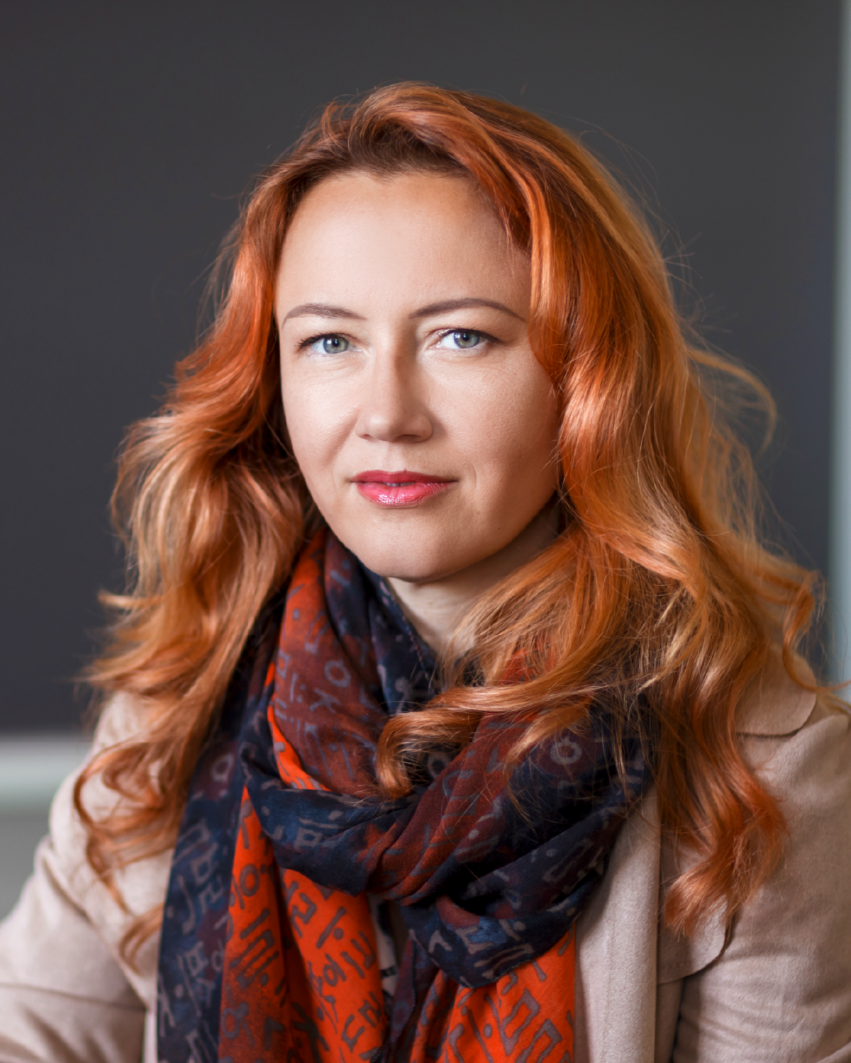 A woman with long red hair wearing a patterned scarf and beige jacket sits in front of a blurred dark background, looking calmly at the camera.