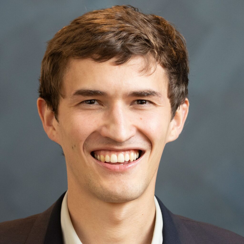 A young man with short brown hair, wearing a dark suit jacket and white shirt, smiles broadly in front of a plain dark gray background.