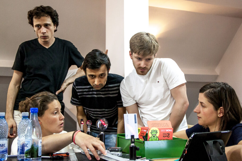 Five people gather around a table with laptops, notebooks, and water bottles, appearing focused and engaged in discussion or problem-solving in a casual indoor setting.