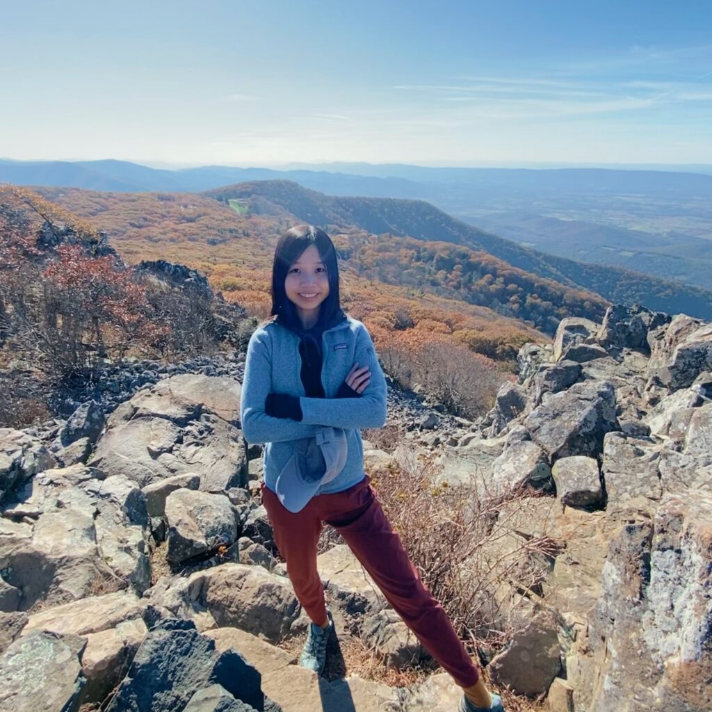 A woman stands on rocky terrain atop a mountain, smiling with arms crossed. Autumn trees and rolling hills stretch into the distance under a clear blue sky.