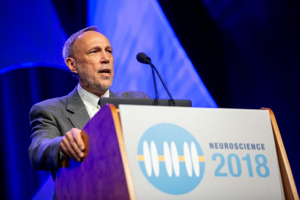 A man in a gray suit speaks at a podium with a microphone. The podium displays a Neuroscience 2018 logo. Blue and white lights are in the background.
