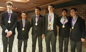 Six young men in suits stand side by side indoors, all wearing gold medals on purple ribbons around their necks. They are smiling and posing for a group photo against a brick wall.