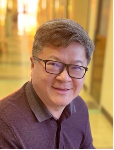 A person wearing glasses and a dark collared shirt smiles at the camera while sitting in a brightly lit hallway with blurred background.