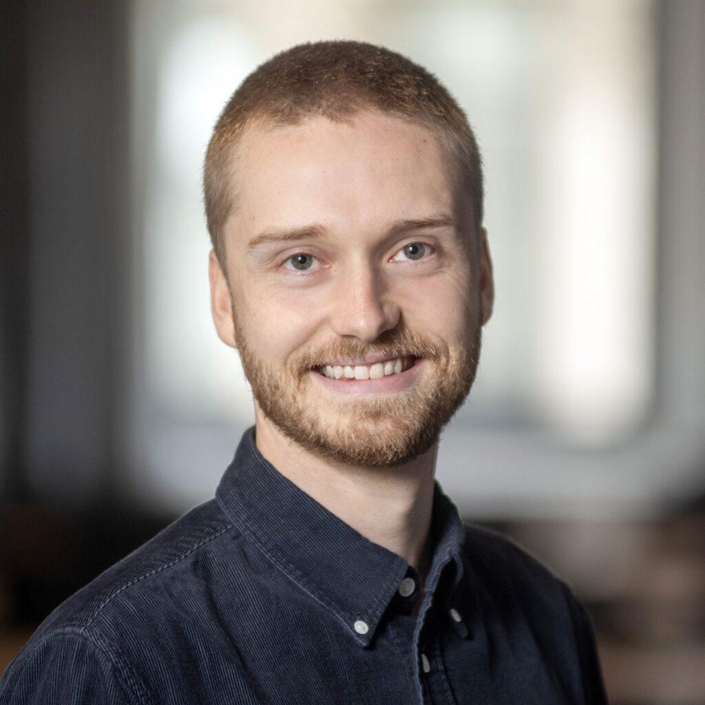 A young man with short hair and a beard, wearing a dark button-up shirt, smiles in a softly lit indoor setting with a blurred background.