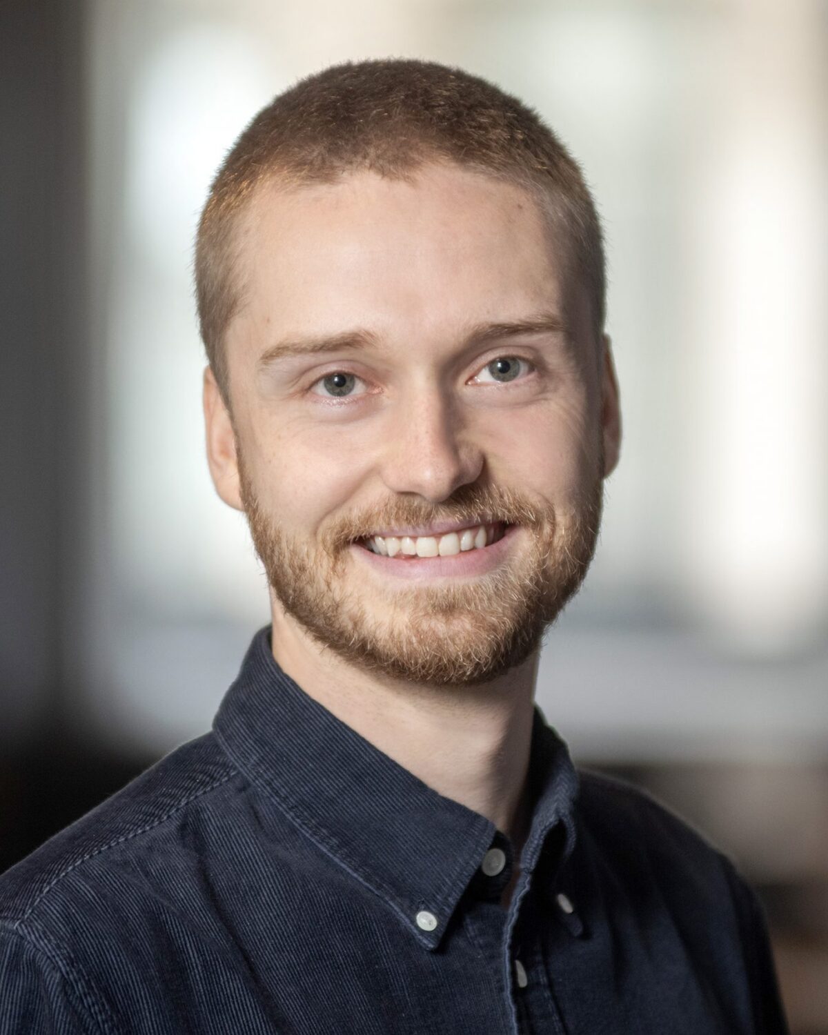 A young man with short hair and a beard, wearing a dark button-up shirt, smiles in a softly lit indoor setting with a blurred background.