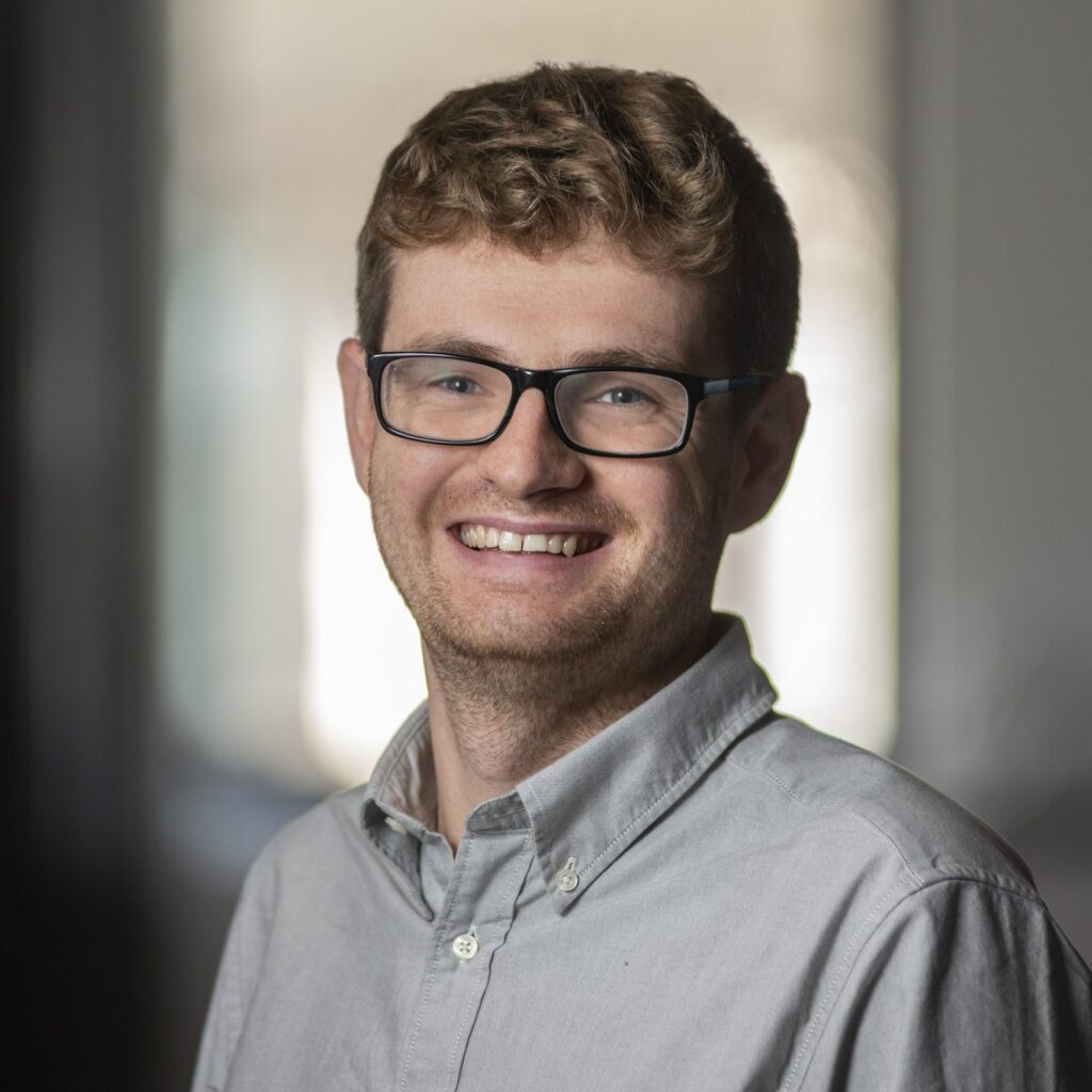 A young man with curly hair, glasses, and a light gray button-up shirt smiles at the camera in a softly lit indoor setting with a blurred background.
