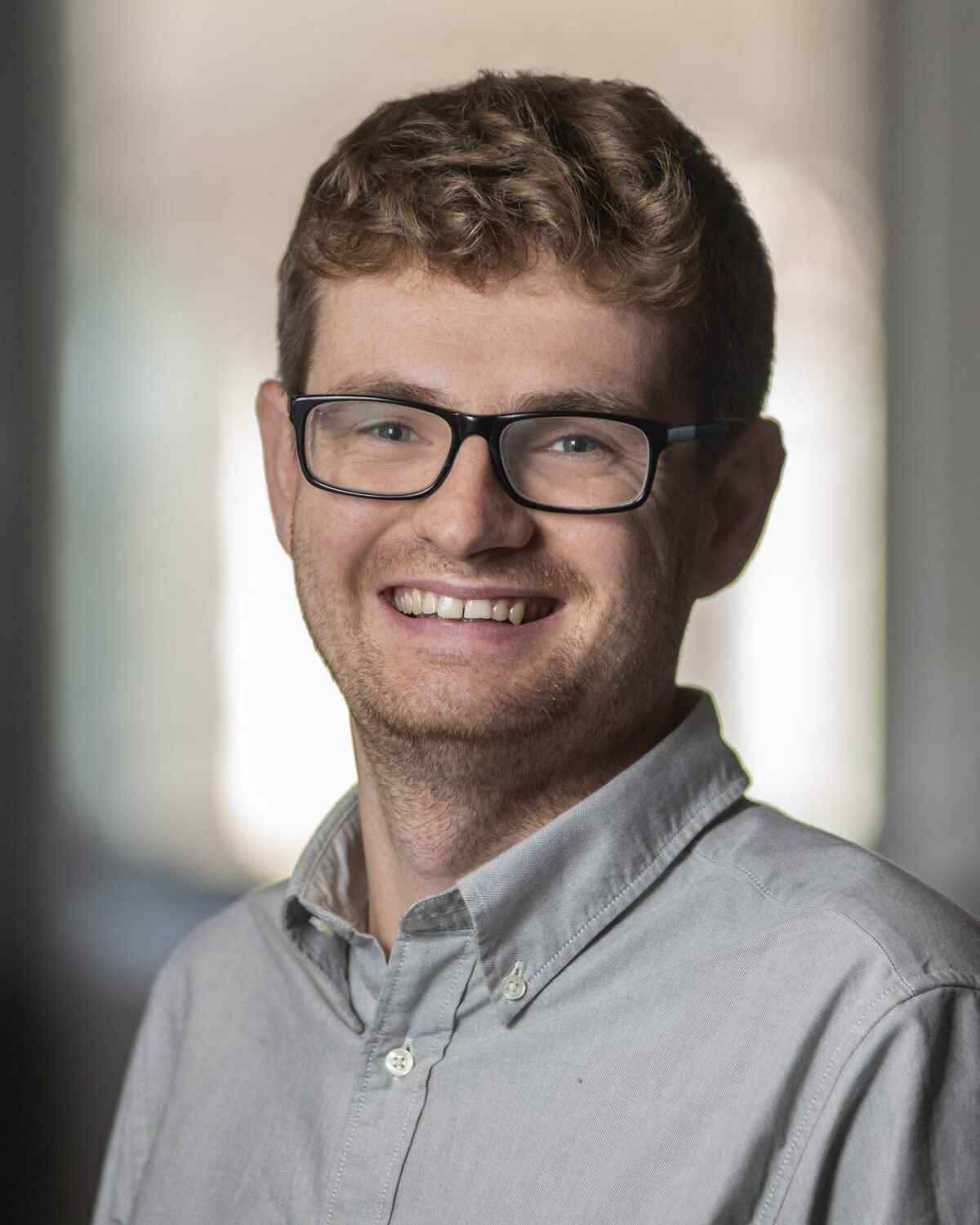 A young man with curly hair, glasses, and a light gray button-up shirt smiles at the camera in a softly lit indoor setting with a blurred background.