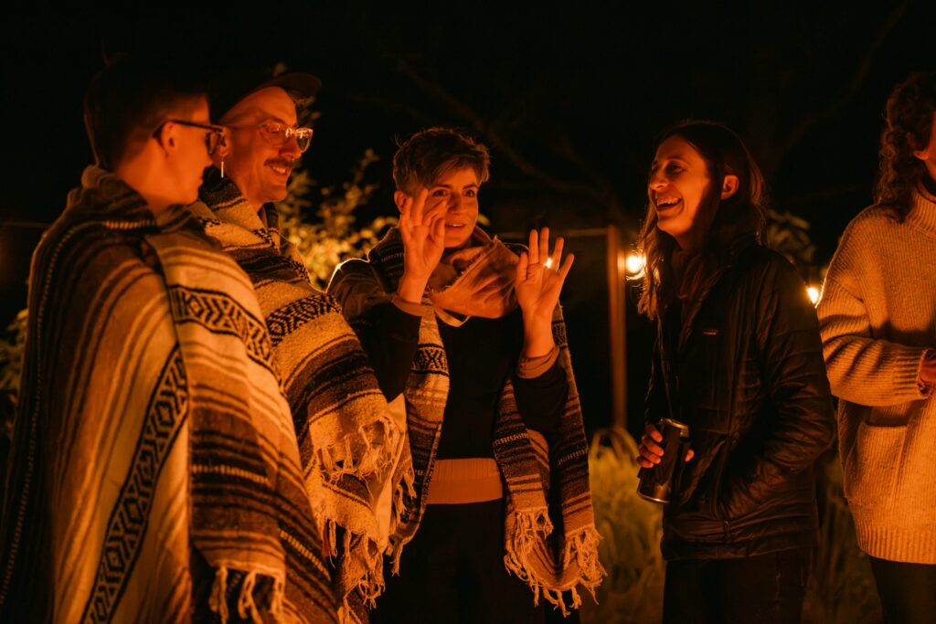 Four people, two wrapped in patterned blankets, smile and chat by a warm outdoor firelight at night. One person gestures with their hands while another holds a drink. The atmosphere is relaxed and friendly.