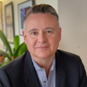 A man with short, neatly styled gray hair, wearing a dark blazer and light blue shirt, sits indoors in front of a plant and framed artwork, looking at the camera with a slight smile.
