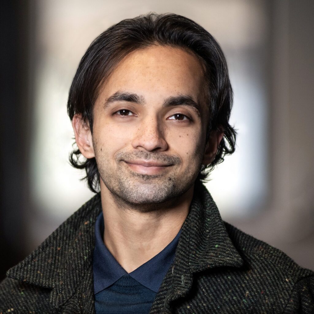 A person with medium-length dark hair and slight facial hair smiles gently, wearing a dark jacket and collared shirt, with a blurred indoor background.