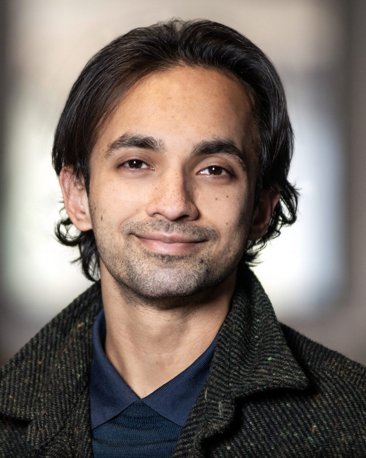 A person with medium-length dark hair and slight facial hair smiles gently, wearing a dark jacket and collared shirt, with a blurred indoor background.