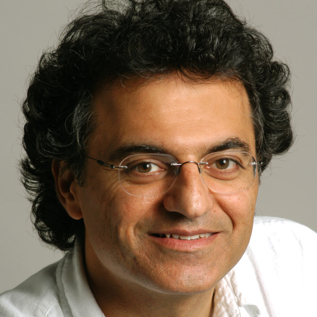 A man with curly dark hair, glasses, and a white shirt is smiling at the camera against a plain light background.