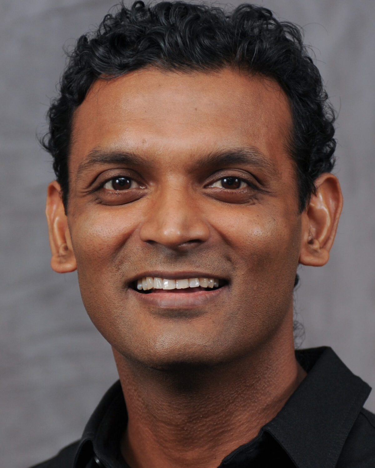 A man with short curly hair and medium brown skin smiles at the camera, wearing a black collared shirt, in front of a plain gray background.