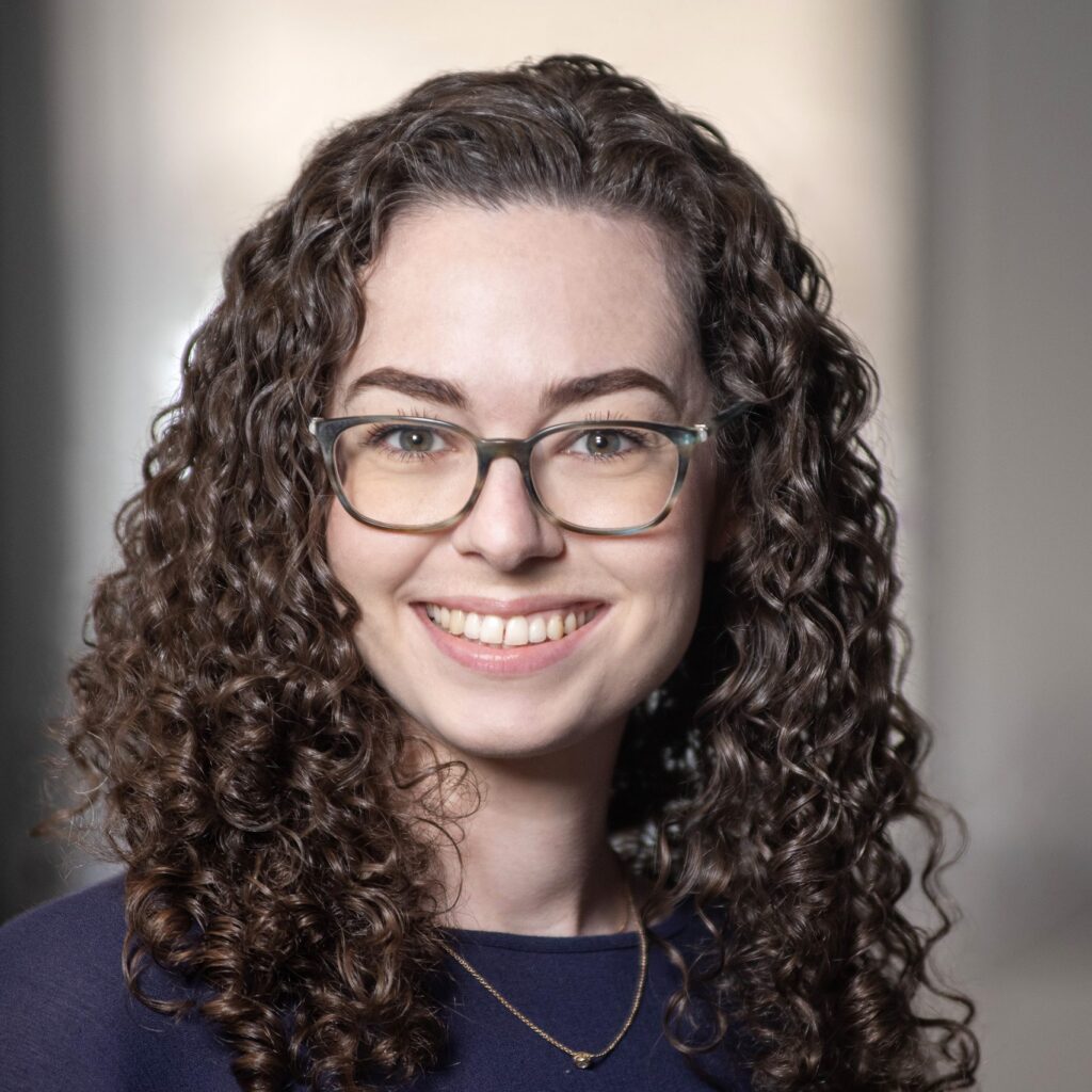 A woman with curly brown hair and glasses smiles at the camera, wearing a dark top and a delicate necklace, with a blurred indoor background.