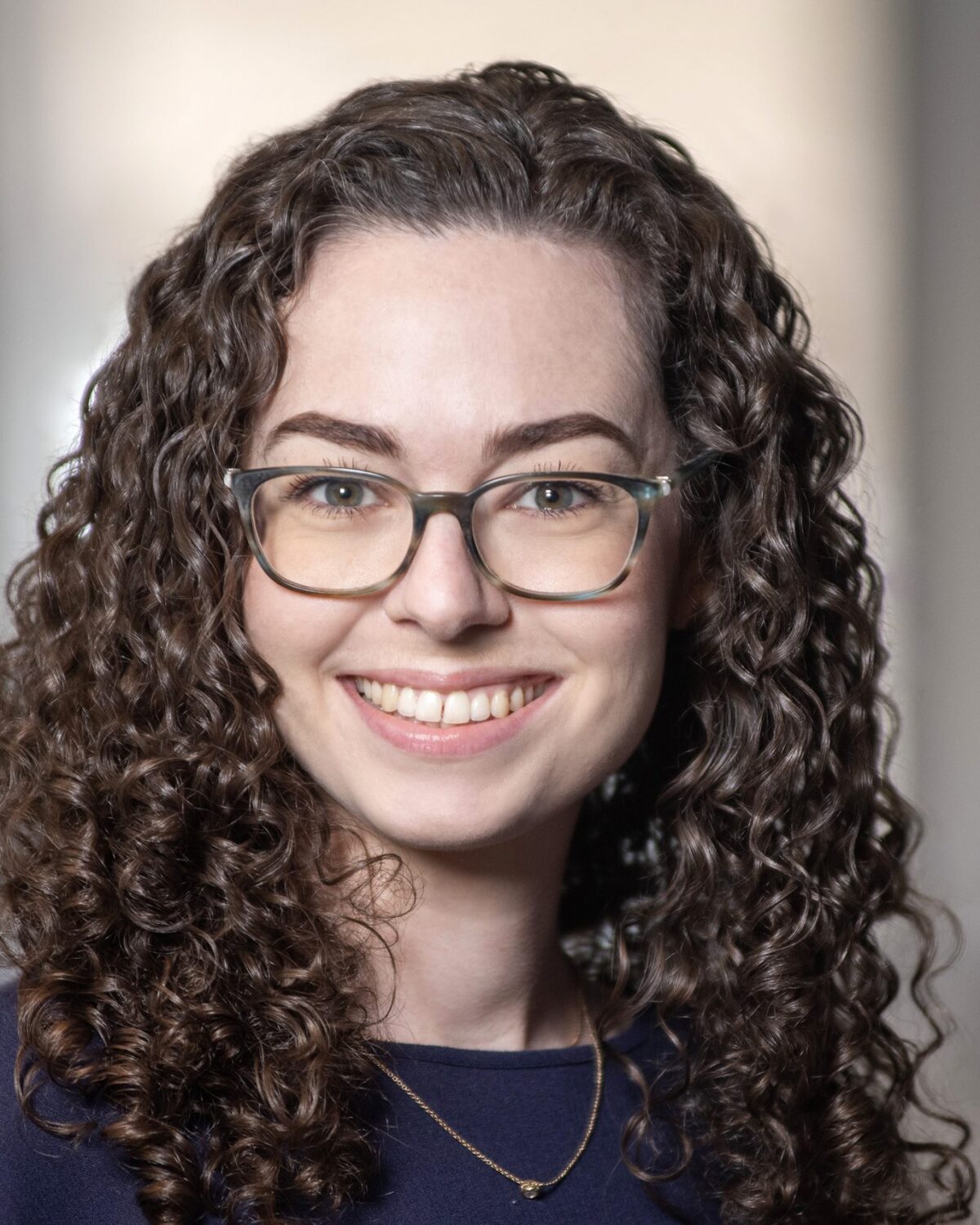 A woman with curly brown hair and glasses smiles at the camera, wearing a dark top and a delicate necklace, with a blurred indoor background.
