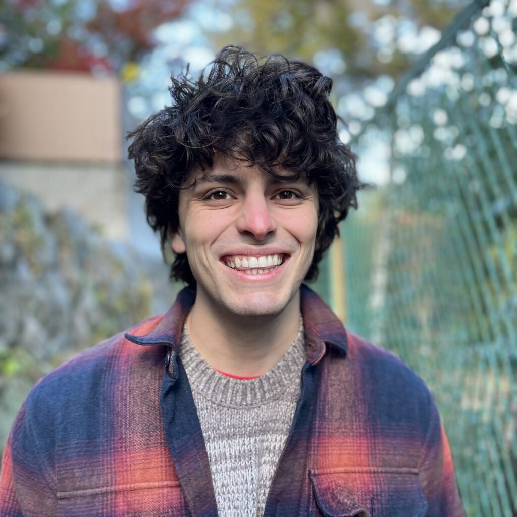 A person with curly dark hair smiles outdoors, wearing a plaid shirt over a knit sweater. A green wire fence and trees with autumn foliage are in the background.