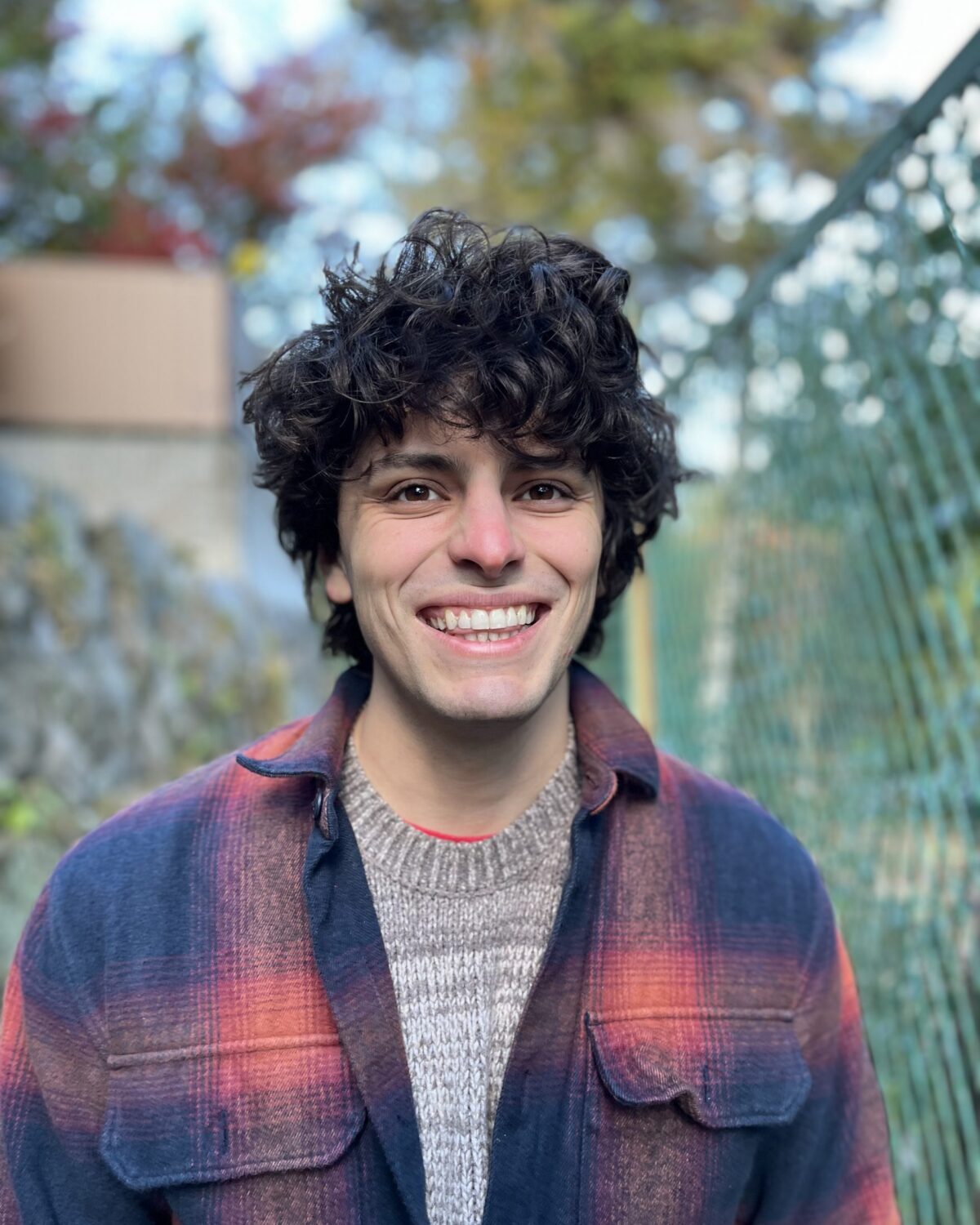 A person with curly dark hair smiles outdoors, wearing a plaid shirt over a knit sweater. A green wire fence and trees with autumn foliage are in the background.